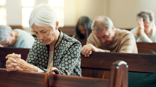 Congregants seated in church pews with heads bowed in prayer, including an elderly woman in the foreground.