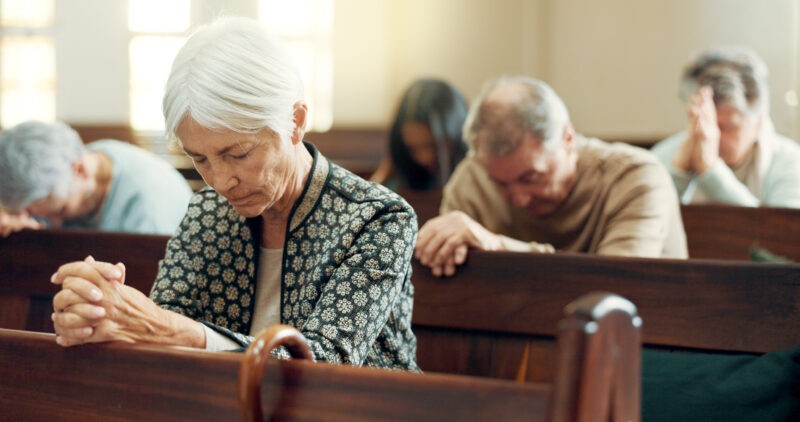 Congregants seated in church pews with heads bowed in prayer, including an elderly woman in the foreground.