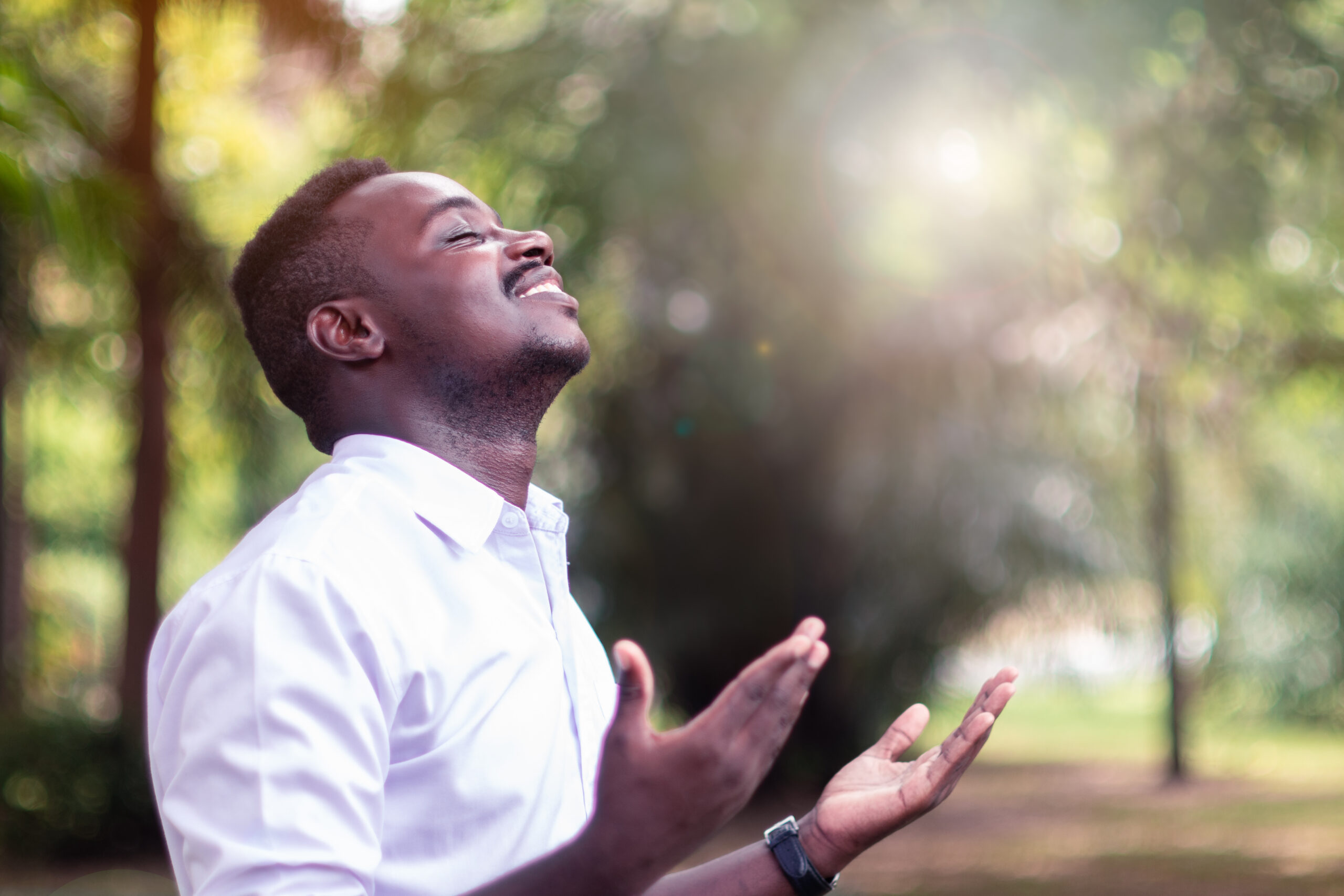 Man standing outdoors with his eyes closed and hands raised in prayer, smiling peacefully in the morning sunlight