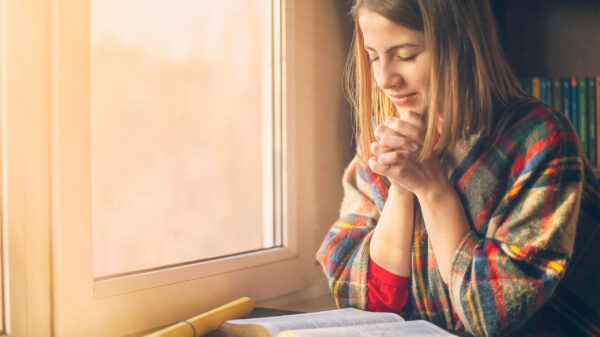 Woman in a cozy plaid sweater praying by the window with an open Bible in front of her, bathed in soft morning light.
