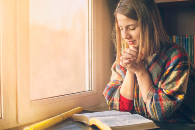 Woman in a cozy plaid sweater praying by the window with an open Bible in front of her, bathed in soft morning light.