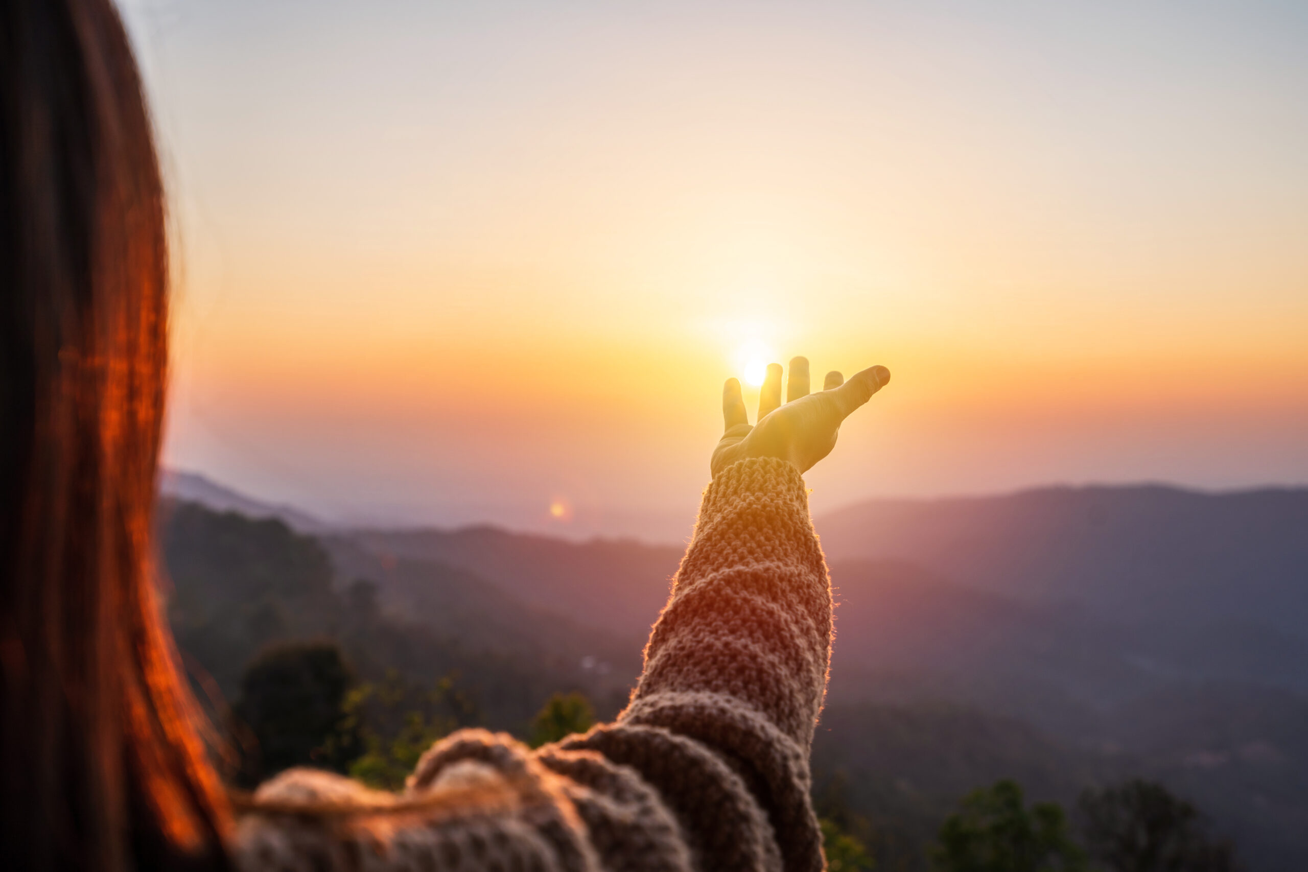 Woman in a cozy sweater reaching her hand toward the rising sun over misty mountains at dawn