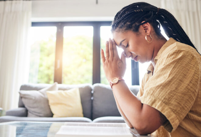 Woman sitting indoors with her head bowed and hands together in prayer, with soft morning light behind her
