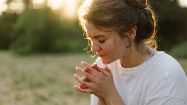 Young woman praying outdoors in soft evening light, hands clasped and eyes closed in peaceful reflection.